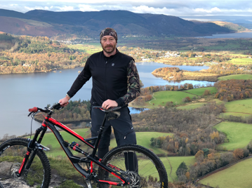 A man with a bike standing in front of a view of a lake and hills.