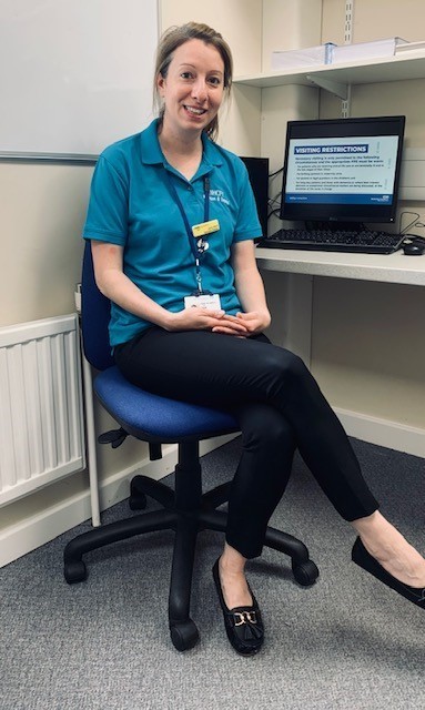 A woman sat a computer. She is wearing a blue polo shirt.