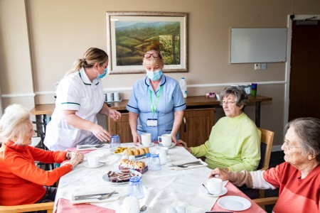 Three elderly women sat around a table being served cups of tea. There are scones on the table.