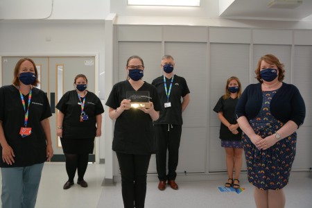 A photograph of six people stood socially distance inside. The woman in the middle is holding an award.