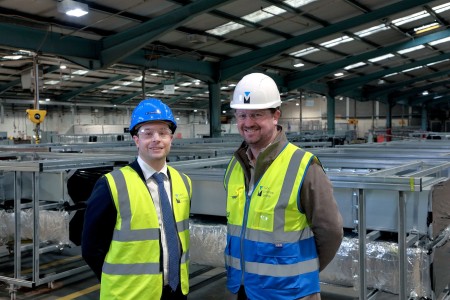 Two men in a factory wearing hi-vis jackets and helmets.