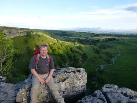 A man sat on a rock that overlooks a view of trees and hills.