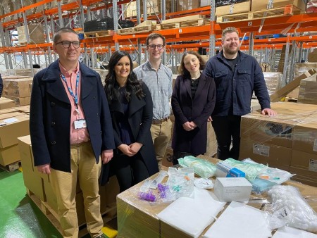 Five people stood together in a warehouse. There are some pieces of medical equipment in front of them.