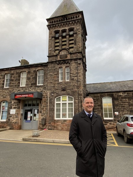 A photograph of a male standing in front of the minor injuries unit at Berwick Infirmary.