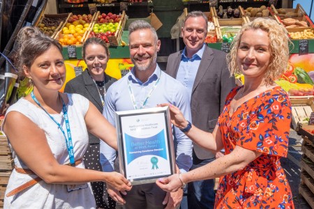 A group of five people stood in front of a fruit and vegetable stall. Two of them are holding an award.