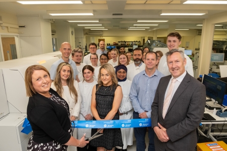 A group of people in the new lab. Someone is at the front cutting a ribbon.