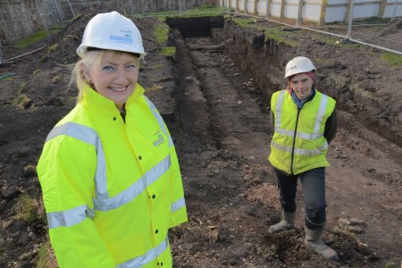 A photograph of a man an a woman in hi-vis jackets and helmets. They are standing where the ground has been dug up.