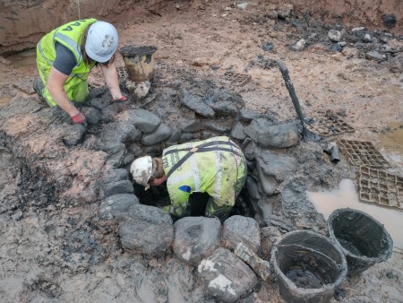 A photograph of two people wearing hi-vis vests and helmets, digging up the ground.