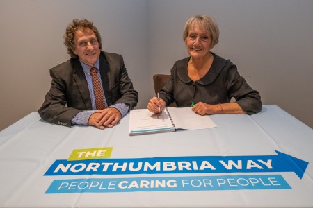 A man and a woman sat at a table. The woman is writing in a book.