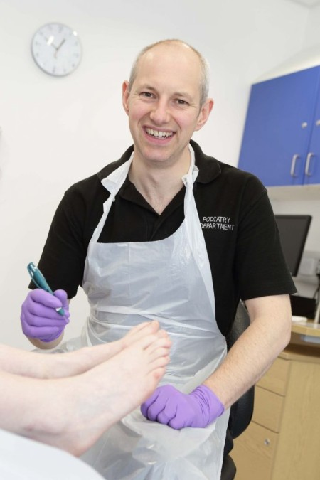 A member of the podiatry department wearing an apron and plastic gloves. A pair of feet from a patient are in front of him.