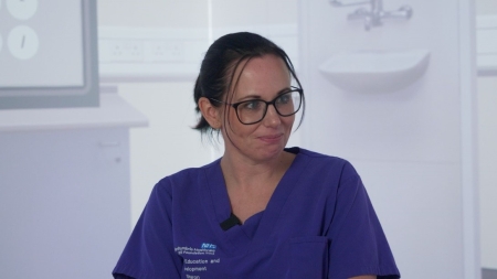 Woman with brunette hair wearing glasses smiling away from the camera wearing purple nurses scrubs