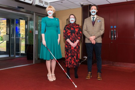 A photograph of two women and a man. They are all wearing facemasks with a clear covering over the mouth. The woman on the left is holding a cane.