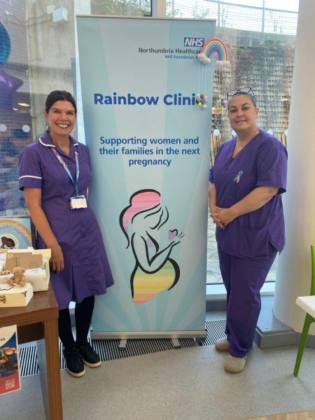 Two midwives in purple uniform standing either side of a rainbow clinic banner