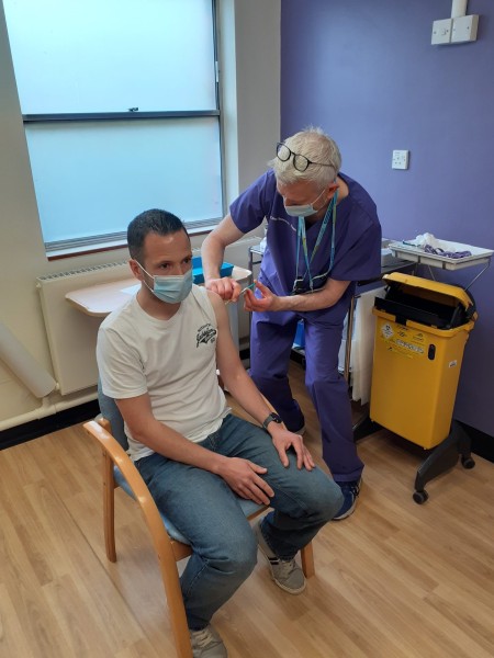 A photograph of a man sat on a chair receiving a vaccination.