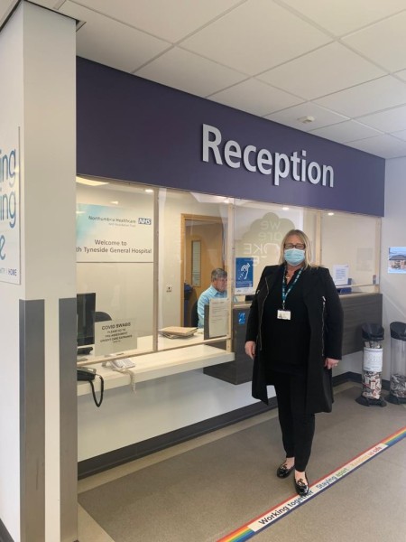 A woman stood at a hospital reception facing the camera. There is a man sat at the reception desk.