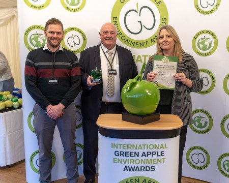 Three people standing behind a Green Apple podium. A man is holding an award and a woman is holding a certificate.