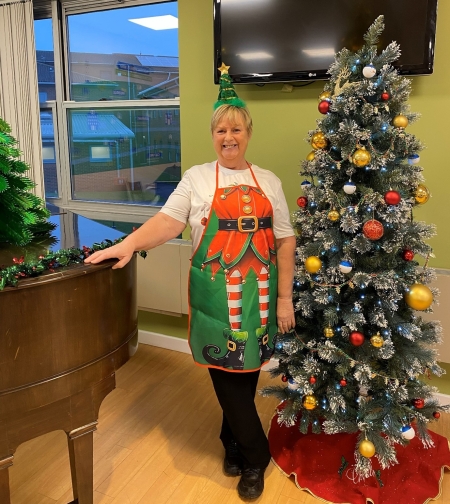 Lady wearing an elf themed apron standing next to a piano and Christmas tree smiling at the camera