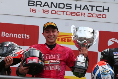 A photograph of a male holding a large, silver trophy. In the other arm he is carrying a helmet.