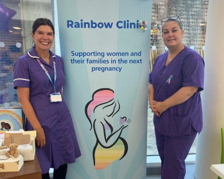Two women standing either side of a blue banner that says Rainbow Clinic.