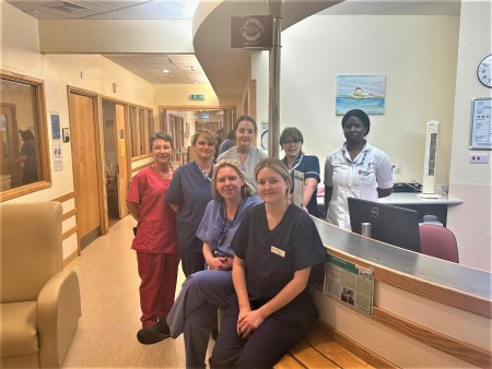 A photograph of seven women in different coloured scrubs and uniforms are standing by the reception of a ward, facing the camera.