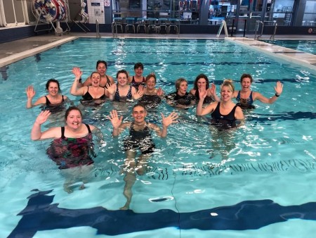 A group of women in a swimming pool. They are all smiling and waving at the camera.