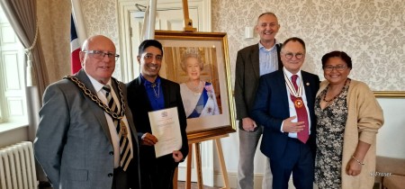 Samridh holding a certificate surrounded by four other people and a photograph of Her Majesty the Queen
