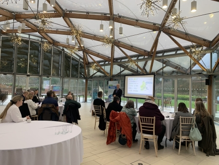 A man presenting at the front of a room with people sat on tables.
