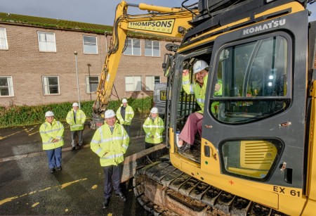 Six people wearing hi-vis jackets and helmets, one of them is sitting in a digger.