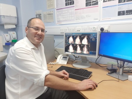 A photograph of a man sat at a desk. On the computer are various scans.