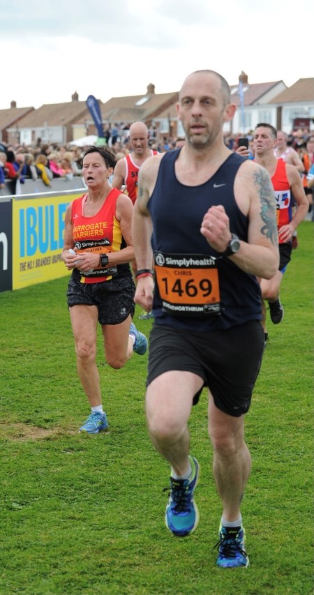 A close up of a man running. There are other people in the background running and a crowd of people watching.