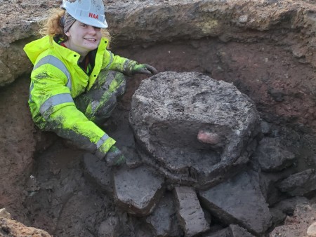 A photograph of a woman wearing a helmet and a hi-vis outfit. She is digging in the ground.