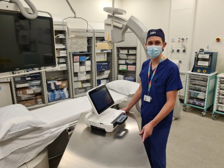 A man wearing scrubs standing in an operating theatre with a computer-like machine in front of him.