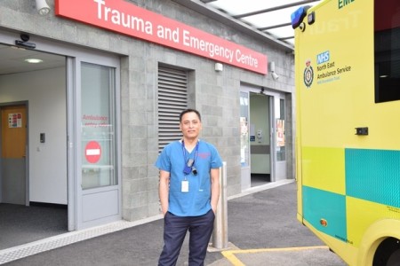 A male is scrubs standing outside a trauma and emergency centre. On the right there is the back end of an ambulance.