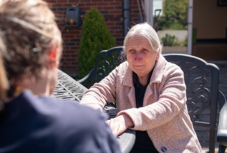 A woman sat in a chair at an outside table looking at someone else.