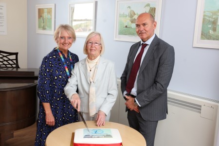 Marion Dickson, Betty Scott and Sir James Mackey stand around table with Betty cutting a celebration cake to celebrate the 30th anniversary of Wansbeck Hospital. 