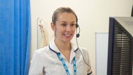 A woman in NHS uniform wearing a headset and looking at a computer.