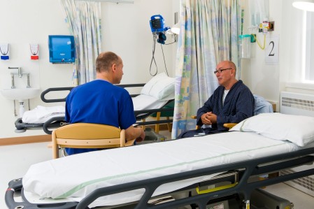 A patient and a doctor sat next to a hospital bed having a conversation.