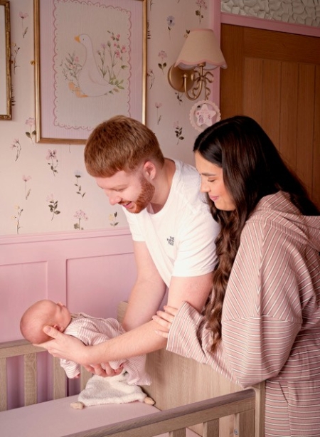 A man with ginger hair and a woman with brown hair holding a baby in a pink cot in a pink decorated room 