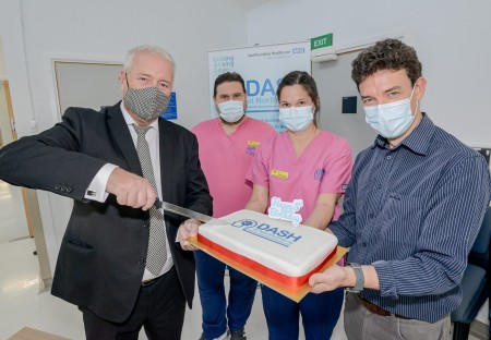 A photograph of four people. Two are wearing pink scrubs. The others are holding a cake while one man cuts into it.