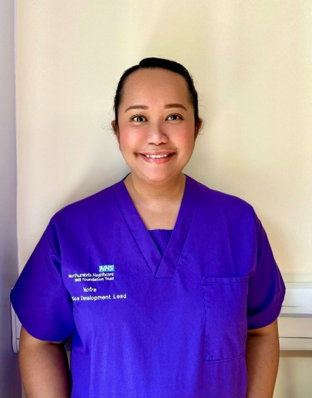 Woman with brunette hair tide up wearing purple scrubs and looking to the camera 