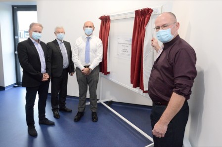 A photograph of four men stood next to a whiteboard. The whiteboard has a pair of red curtains that have been opened.